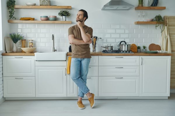 Full length of thoughtful young man leaning at the kitchen desk looking away